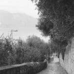 Analoges Schwarz-Weiß Foto zeigt einen alten gepflasterten Weg, der einen Berg hinab führt, im Hintergrund ist der Gardasee zu sehen