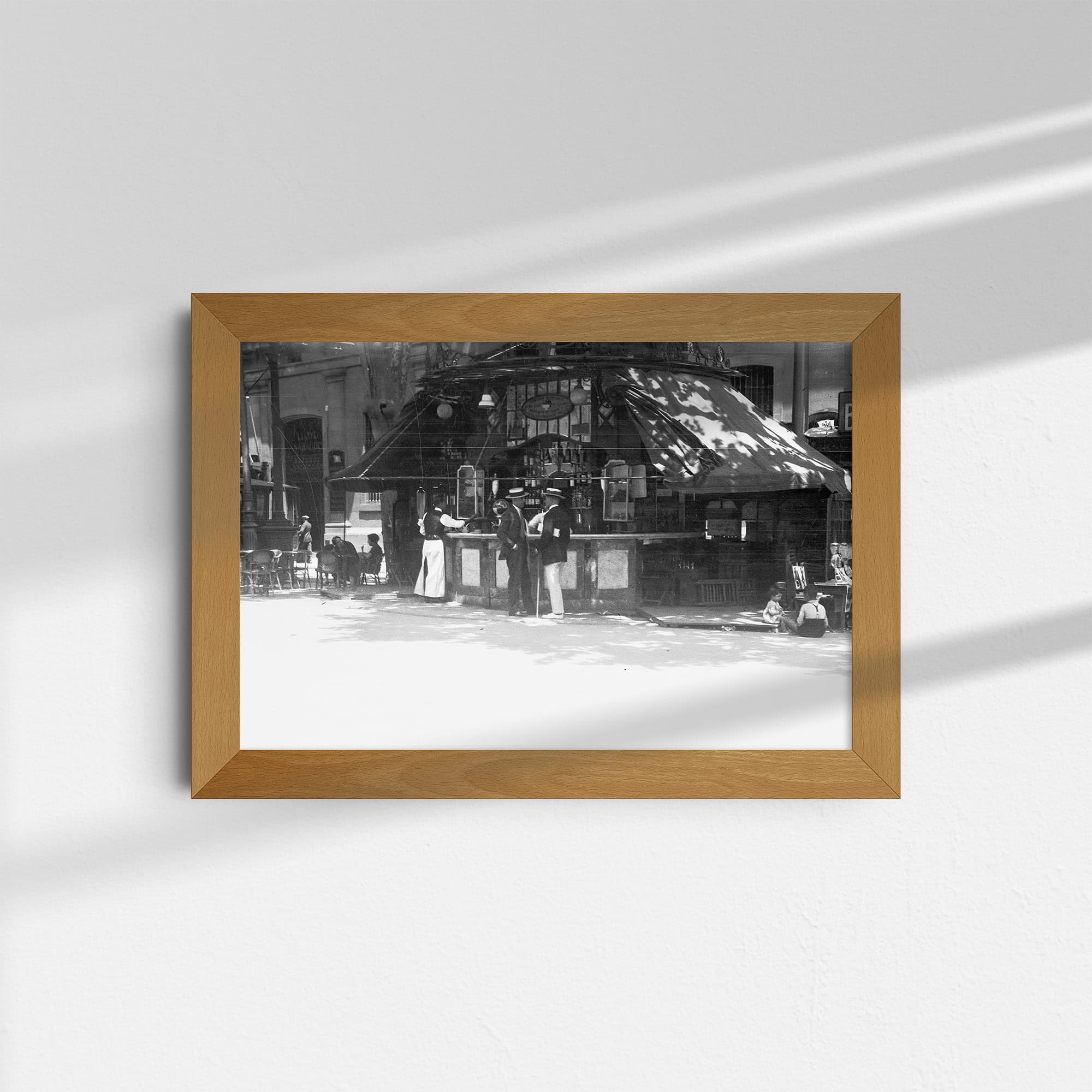 Dieses Schwarz-Weiß Bild zeigt einen Ausschnitt der berühmten Promenade La Rambla in Barcelona, Spanien. Vor einem für diese Straße sehr typischen Kiosk mit Getränken stehen zwei Männer in der typischen Kleidung der 20er Jahre und unterhalten sich. Auf der rechten Seite des Kiosk sitzen Kinder am Boden, auf der linken Seite sind Tische und Stühle für Gäste zu sehen. Das Foto wurde um das Jahr 1930 aufgenommen.