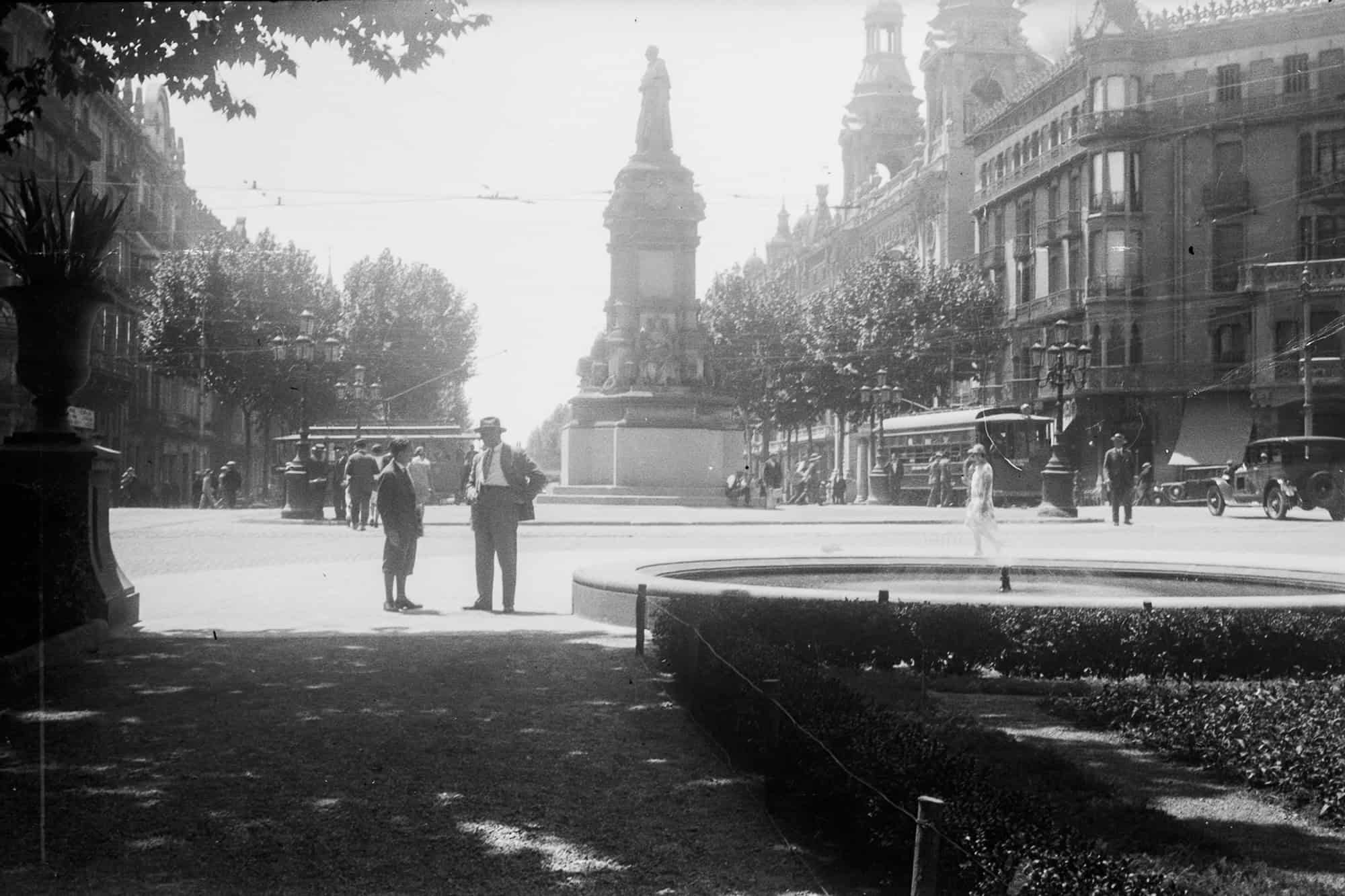 Dieses alte Schwarz-Weiß Foto zeigt eine Straßenszene in Barcelona um das Jahr 1930. Zu sehen ist die Kreuzung Gran Vía und Rambla de Catalunya mit dem alten Denkmal für Joan Güell i Ferrer, das im Jahr 1936 zerstört wurde. Im Vordergrund unterhalten sich zwei Männer. Es sind alte Straßenbahnen und Autos zu sehen.