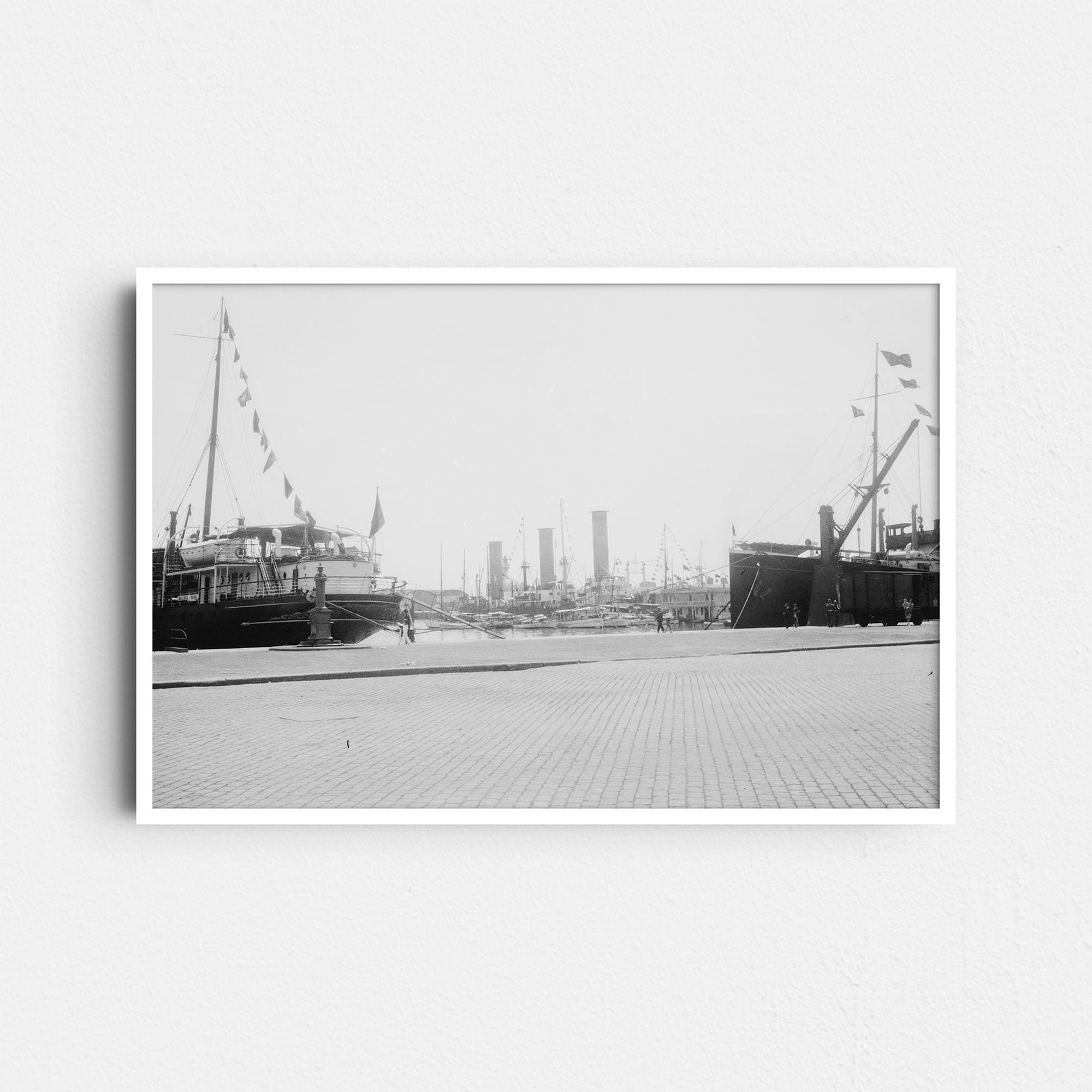 Dieses historische Schwarz-Weiß Foto zeigt eine Szene aus dem Hafen in Barcelona um das Jahr 1930. Am linken und am rechten Rand sind Bug bzw. das Heck zweier großer Dampfschiffe zu sehen. An dem rechten Schiff sind zusätzlich ein Ladekran und ein Güterwaggon zu sehen. Im Zentrum zwischen den beiden Schiffe sind zahlreiche kleinere Boote und Hafengebäude zu erkennen.