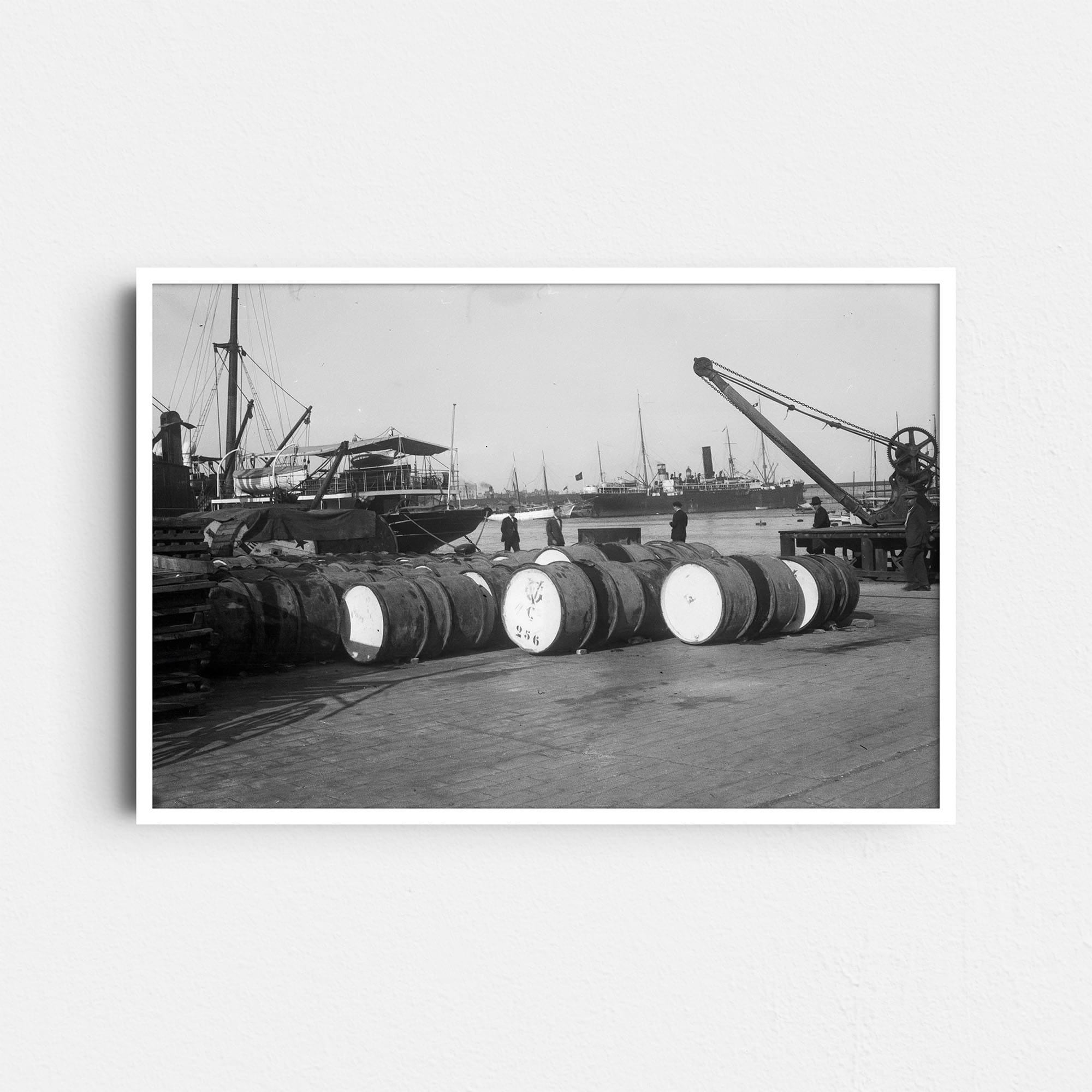 Dieses alte Schwarz-Weiß Foto zeigt eine Szene aus einem großen Hafen. Im Vordergrund lagern Güter auf dem Kai, im Hintergrund sind Schiffe und ein Kran zu sehen. Die Fotografie wurde um das Jahr 1930 in Barcelona aufgenommen.