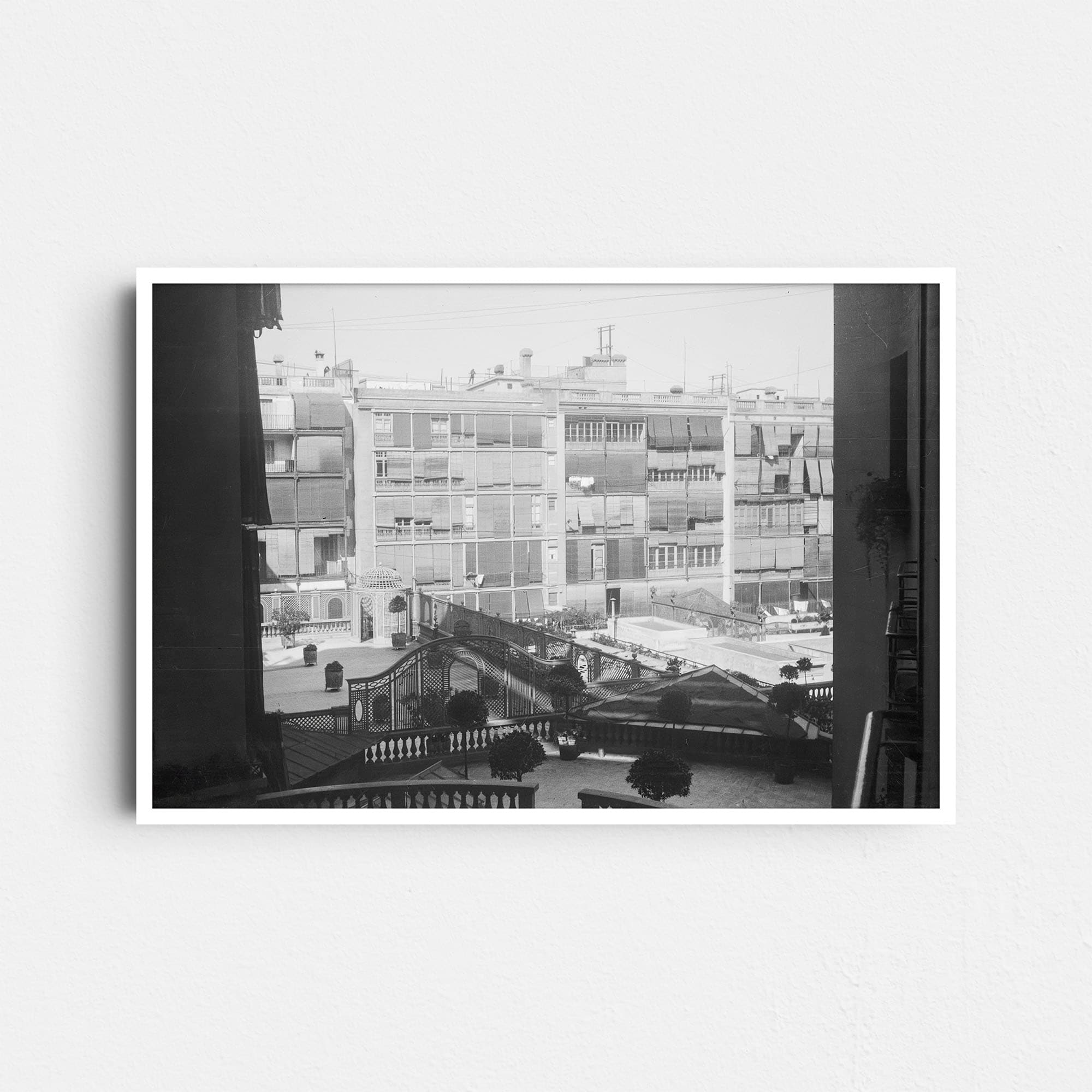 Diese Schwarz-Weiß Fotografie zeigt ein recht typisches Bild städtischer Architektur in Spanien. Es zeigt einen Innenhof mit einer Terrasse mit einem kunstvollen Metallgeländer und kleinen Bäumen in Kübeln. Dahinter sind die Fassaden von Wohngebäuden zu sehen. Zur Schutz vor der Sonne sind Balkone und Fenster mit Jalousien verdunkelt. Diese Aufnahme zeigt den Innenhof des ehemaligen Hotel Ritz in Barcelona, das heute "El Palace" heißt, um das Jahr 1930.