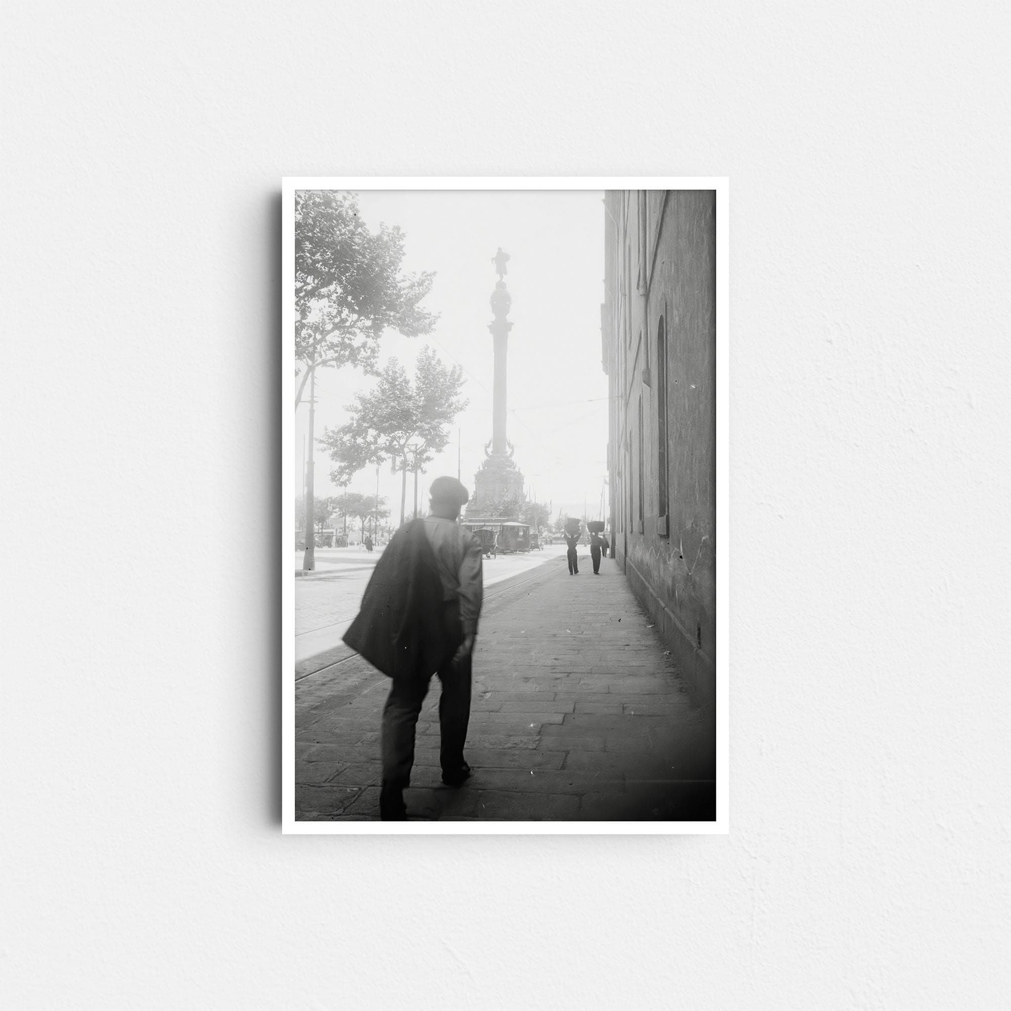Dieses historische Foto zeigt eine Straßenszene in Barcelona, Spanien. Im Vordergrund ist ein Gehweg an einer Straße, auf dem ein Mann geht. Rechts ist die Häuserwand eines Gebäudes zu sehen. Im Hintergrund ist das Kolumbus-Denkmal (Monumento a Colón) zu erkennen. Am Fuß der Säule sind eine Straßenbahn, ein Pferdefuhrwerk und ein Automobil zu sehen. Das Bild um das Jahr 1930 aufgenommen.