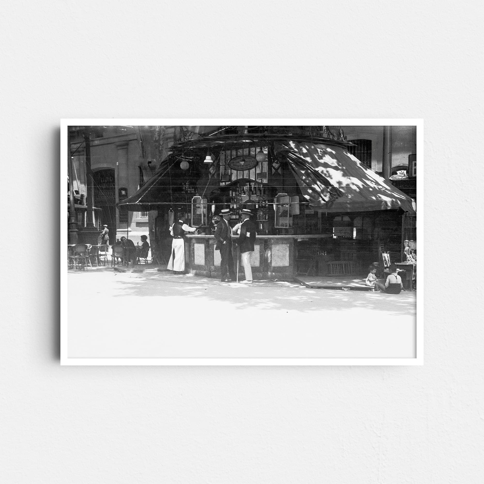 Dieses Schwarz-Weiß Bild zeigt einen Ausschnitt der berühmten Promenade La Rambla in Barcelona, Spanien. Vor einem für diese Straße sehr typischen Kiosk mit Getränken stehen zwei Männer in der typischen Kleidung der 20er Jahre und unterhalten sich. Auf der rechten Seite des Kiosk sitzen Kinder am Boden, auf der linken Seite sind Tische und Stühle für Gäste zu sehen. Das Foto wurde um das Jahr 1930 aufgenommen.