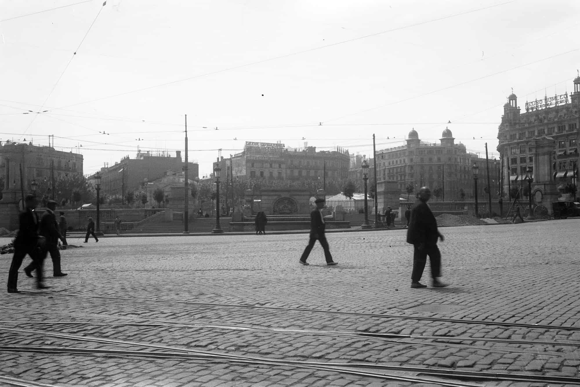Dieses historische Schwarz-Weiß Foto zeigt die Plaça de Catalunya in Barcelona. Dieser Platz war um 1930 ein wichtiger Verkehrsknotenpunkt, was auch an den vielen Straßenbahnschienen und Oberleitungen zu sehen ist. Im Vordergrund überqueren Männer den Platz. Im Zentrum ist die Gestaltung des Platzes zur Wertausstellung 1929 zu sehen, die zum Zeitpunkt der Aufnahme noch nicht ganz fertig ist. Im Hintergrund sind die Gebäude am Rand des Platzes zu sehen.