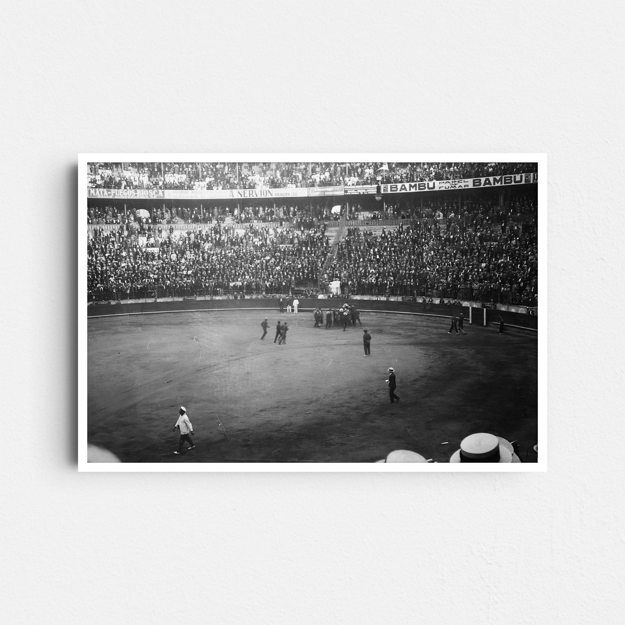 Dieses historische Schwarz-Weiß Foto zeigt einen Ausschnitt des Innenraumes einer Stierkampfarena in Barcelona, Spanien, um das Jahr 1930. Im Zentrum ist die Arena mit einer Ansammlung von Menschen zu sehen. Die Zuschauerränge sind voll besetzt. An den Rängen ist Werbung zu erkennen.