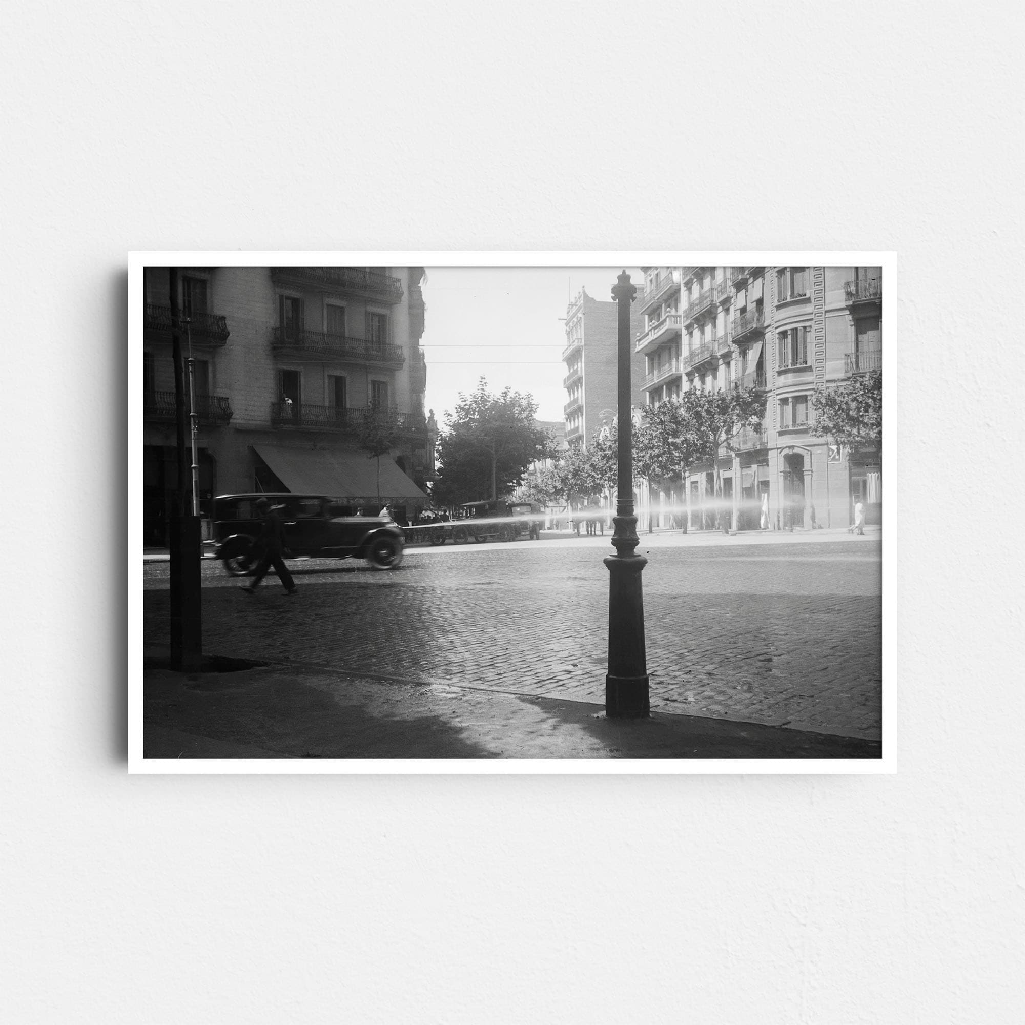 Diese Schwarz-Weiß Fotografie zeigt eine Straßenkreuzung in Barcelona um das Jahr 1930. Ein Mann reinigt die Straße mit einem Wasserschlauch, während ein Auto an ihm vorbeifährt. Es handelt sich um ein historisches Foto, das das Stadtbild und den Verkehr einer vergangenen Epoche dokumentiert.