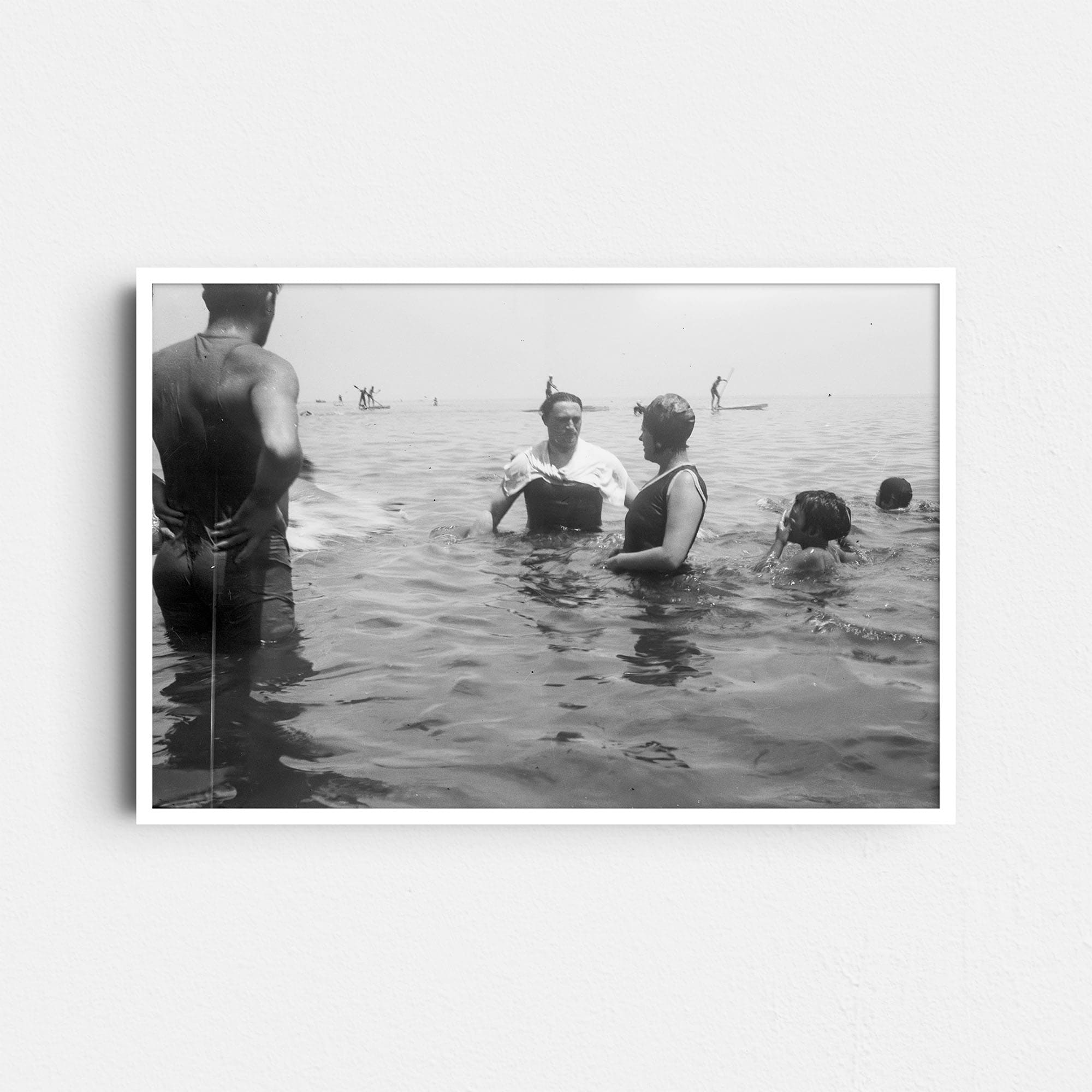 Dieses historische Schwarz-Weiß Foto zeigt im Meer Badende um das Jahr 1930. Im Zentrum sind ein Mann und eine Frau in der damals typischen Badebekleidung zu sehen, neben denen Kinder im Wasser schwimmen. Im Hintergrund sind mehrere Menschen auf Stand-up-Paddling Boards zu sehen.