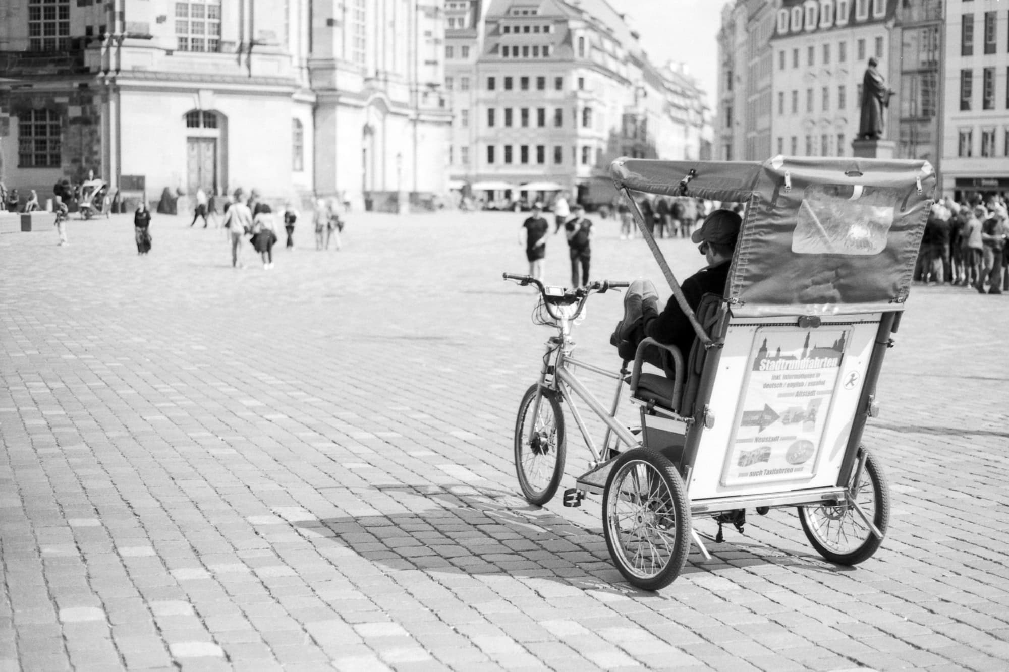 Diese Schwarz-Weiß Fotografie wurde am Neumarkt in Dresden aufgenommen und zeigt einen Blick in Richtung Frauenkirche. Im Vordergrund wartet eine Fahrradrikscha auf Kundschaft.