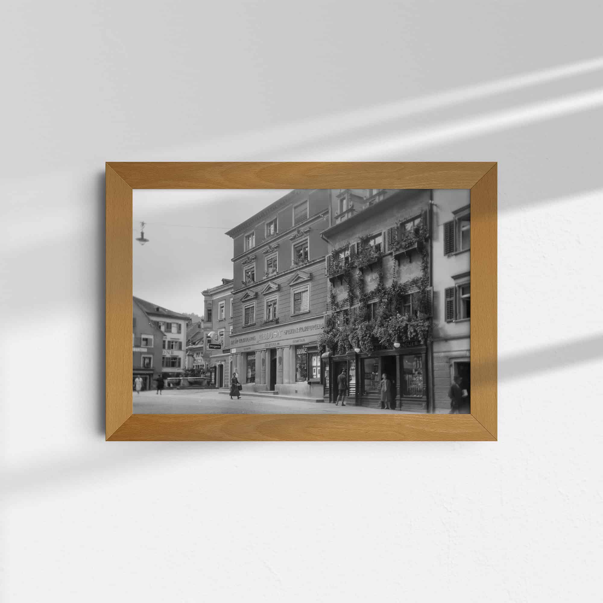 Diese historische Schwarz-Weiß Fotografie zeigt einen Teil der Rathausstraße in Bregenz, Österreich, um das Jahr 1930. Im Zentrum des Bildes stehen zwei alte Häuser. Das Haus in der Mitte beherbergt die Parfümerie Medusa, die noch heute existiert. Das kleinere Haus rechts daneben ist dicht bewachsen. Vor dem Geschäft in diesem Haus steht eine Frau und schaut zum Fotografen.