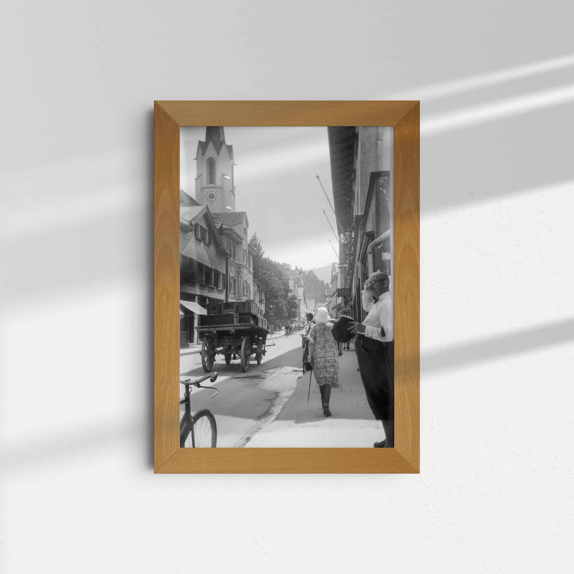 Diese historische Schwarz-Weiß Fotografie wurde um 1930 in der Ludwigstraße in Garmisch-Partenkirchen aufgenommen. Es zeigt den Straßenverlauf in Richtung der Pfarrkirche Maria Himmelfahrt, deren Turm auf der linkte im Hintergrund erkennbar ist. Links und rechts der Straße befinden sich alte Häuser. Auf der Straße ist ein Pferdefuhrwerk zu sehen, auf dem Gehweg laufen Passanten. Am rechten Bildrand steht ein Mann und beobachtet die Szenerie.