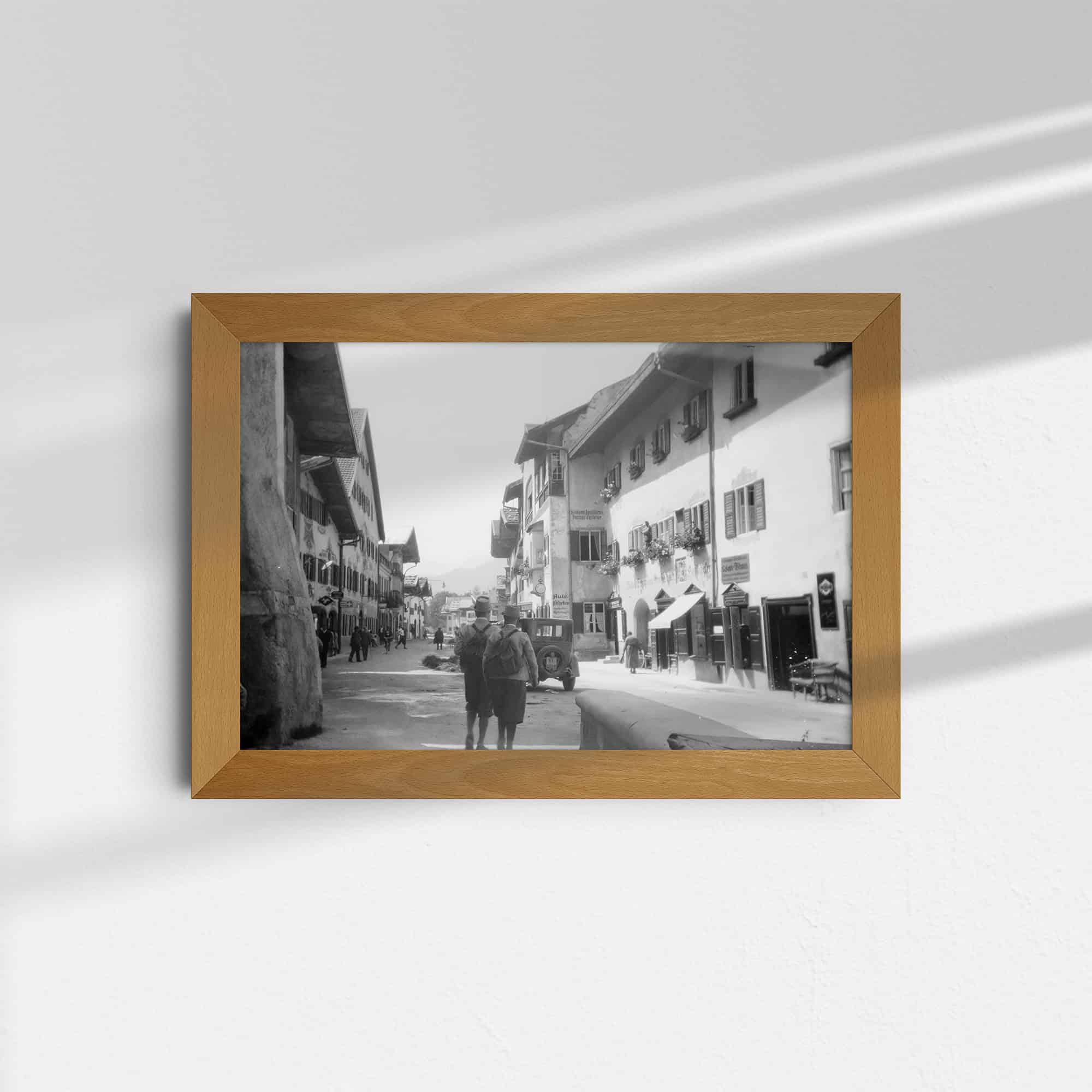 Dieses Foto zeigt eine historische Ansicht der Marktstraße in Mittenwald, Deutschland, um das Jahr 1930. Der Blick geht die Straße entlang, im Hintergrund sind Berge zu sehen. Links und rechts der Straße sind Häuser in der typischen bayerischen Architektur mit verzierten Fassaden zu sehen, die für diese Region charakteristisch sind. Auf der Straße sind Passanten und ein altes Auto zu sehen.