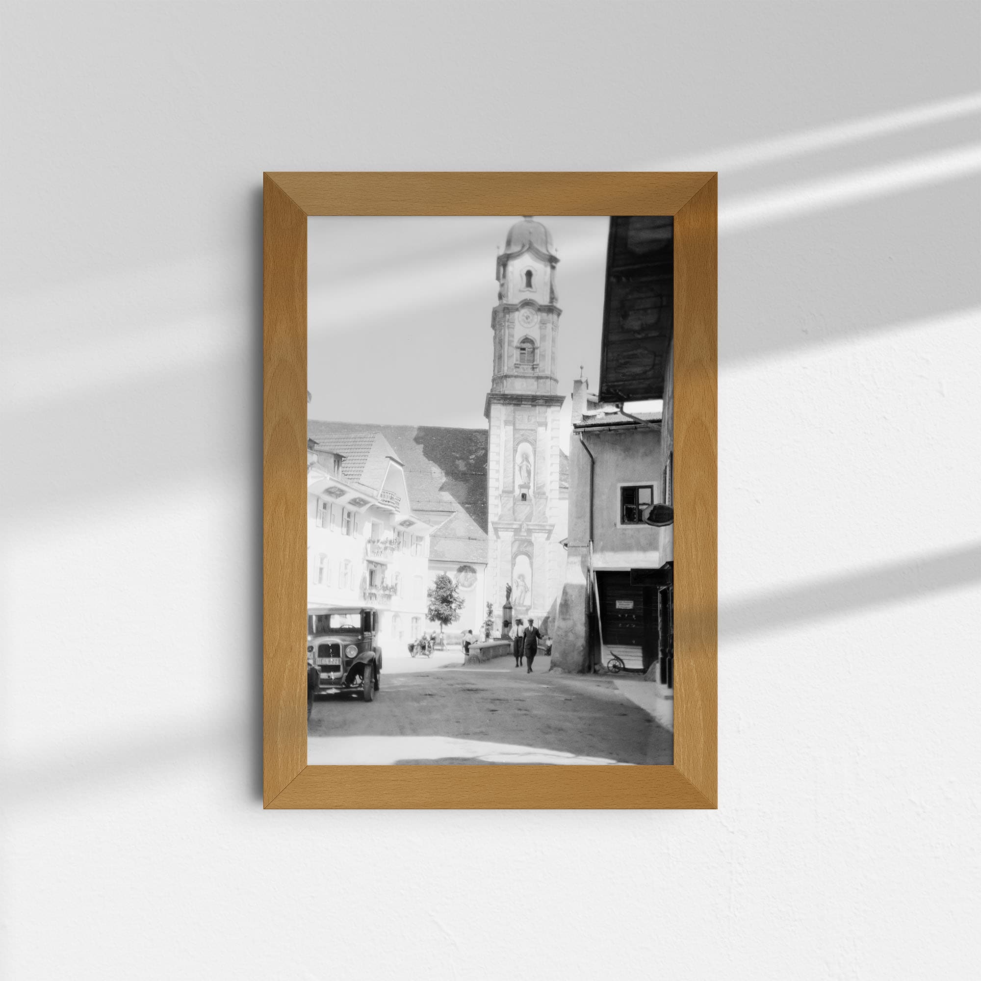 Dieses um das Jahr 1930 aufgenommene Schwarz-Weiß Foto zeigt die mit Fresken geschmückte Pfarrkirche St. Peter und Paul in dem Ort Mittenwald in Bayern, Deutschland. Im Zentrum des Bildes steht der Turm der Kirche. Links und rechts sind Häuser des Ortes zu sehen. Am rechten Rand steht ein altes Auto, auf der Straße sind Passanten unterwegs.