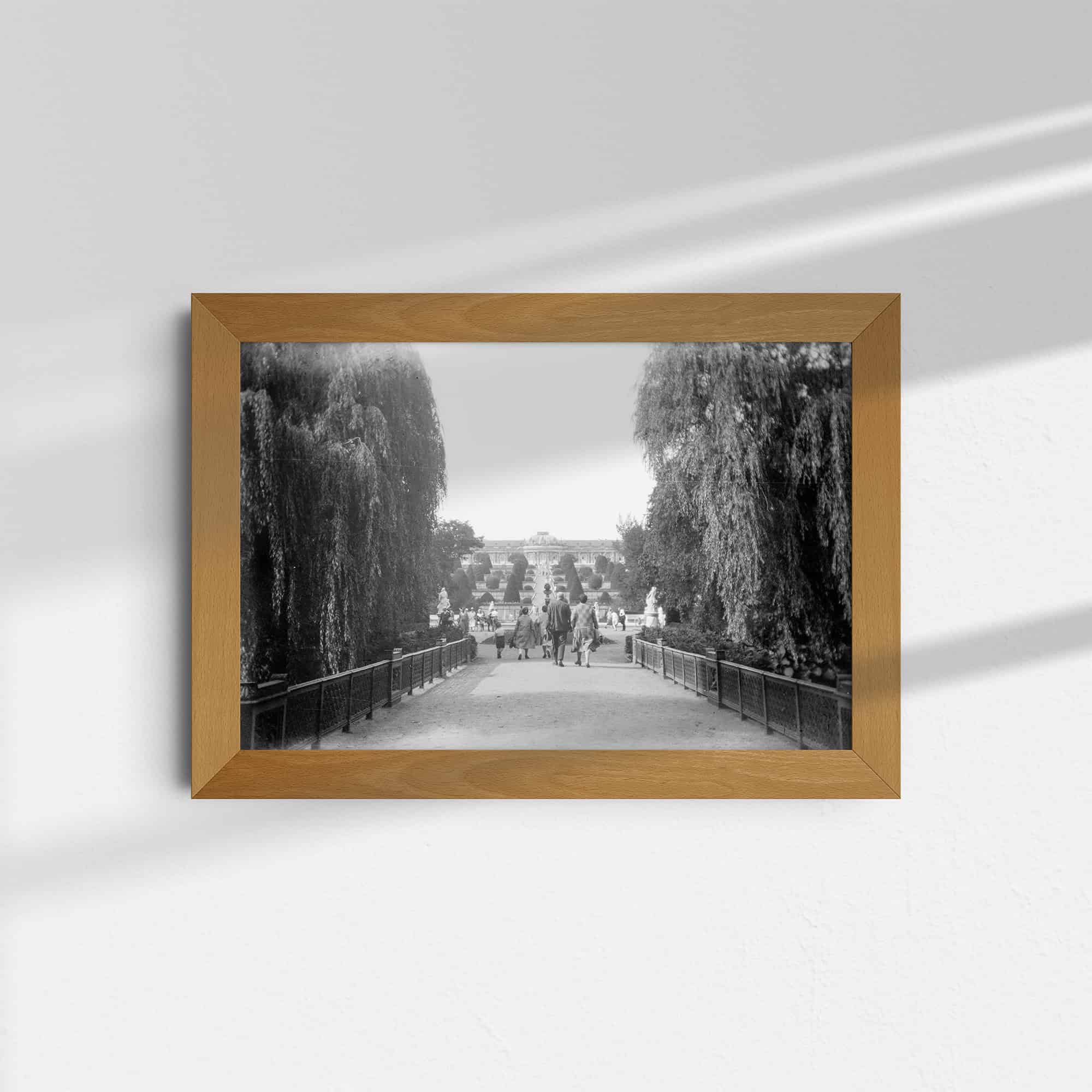 Dieses historische Foto zeigt den Park Sanssouci in Potsdam, Deutschland, im Jahr 1929. Links und rechts stehen dicht belaubte Bäume. Im Zentrum des Bildes ist im Hintergrund das auf einem Hügel gelegene Schloss Sanssouci zu sehen. Davor sind Spaziergänger zu sehen.