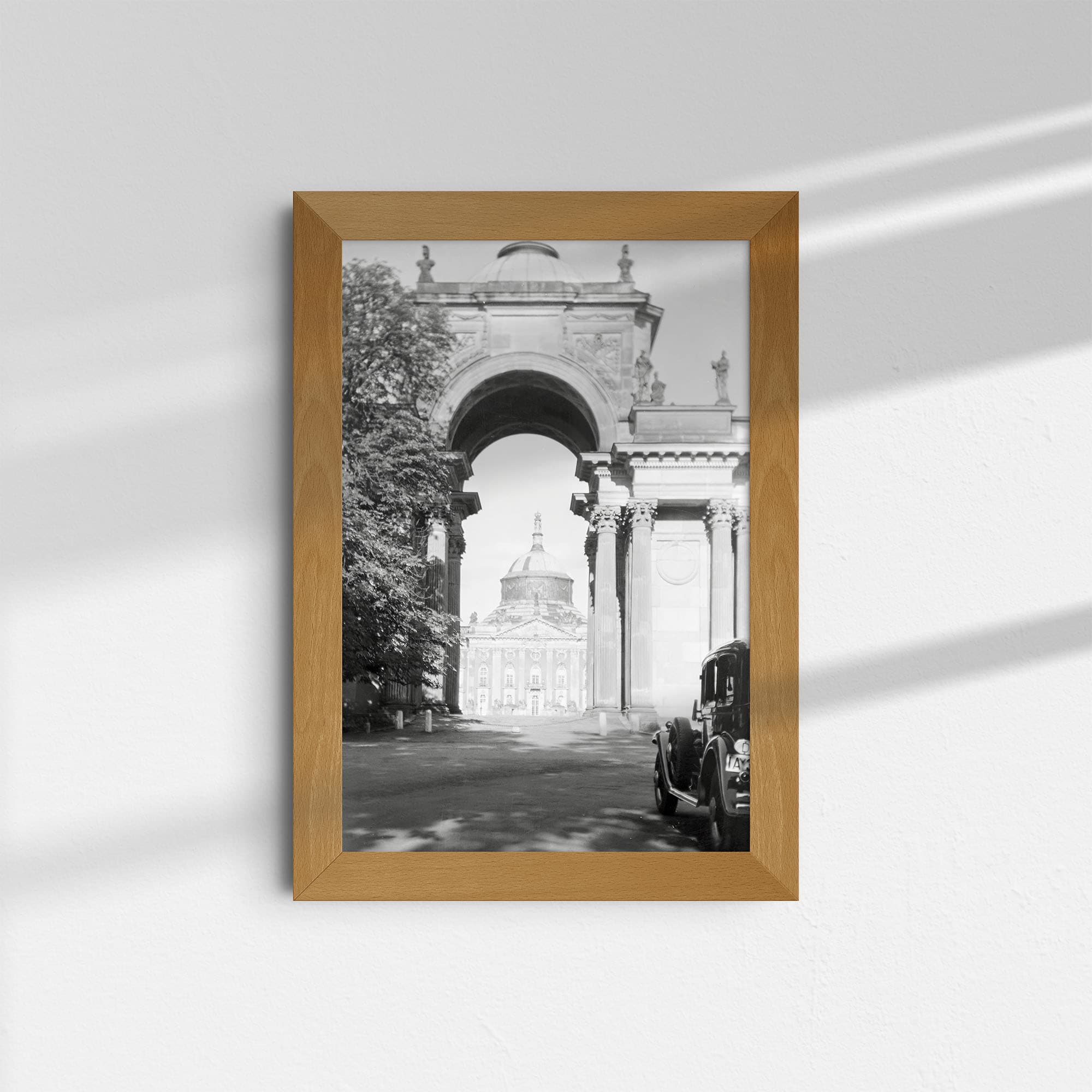 Dieses historische Schwarz-Weiß Foto zeigt den Blick durch das Triumphtor auf das Neue Palais im Park Sanssouci in Potsdam im Jahr 1929. Der hohe Bogen des Tores steht im Zentrum des Bildes. In dem Bogen ist der Mittelteil des Neuen Palais zu erkennen. Links neben dem Tor steht ein Baum. Am rechten Bildrand ist ein Automobil dieser Zeit zu sehen.