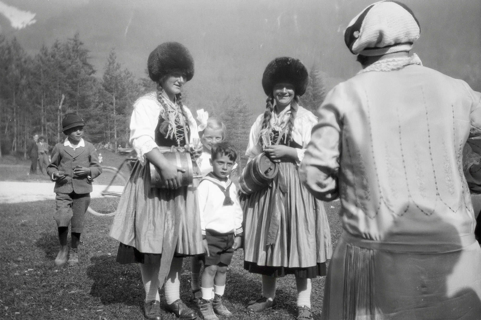Dieses historische Schwarz-Weiß Foto zeigt zwei Frauen in Tirol in der historischen Tracht der Marketenderinnen mit der charakteristischen Fellmütze. Beide haben ein kleines Holzfass umgehängt, in dem sich Schnaps befindet. Neben ihnen stehen Kinder. Recht ist eine Frau in der typischen Kleidung der 1920er Jahre zu sehen.