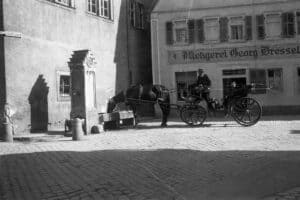 Dieses alte Schwarz-Weiß Foto zeigt eine Straßenszene, die in Heidingsfeld bei Würzburg, Deutschland, um das Jahr 1930 aufgenommen wurde. Eine von einem Pferd gezogene Kutsche hält an dem historischen Brunnen am Rathaus, um das Tier trinken zu lassen.