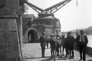 Dieses historische Foto zeigt den Alten Kranen in Würzburg, einen barocken Hafenkran aus dem Jahr 1773 am Mainufer. Es handelt sich um einen Steintretkran mit Doppelausleger, der ursprünglich durch Menschenkraft im Inneren betrieben wurde. Die Szene zeigt Personen in historischer Kleidung und einen Esel vor der Anlage, die heute ein geschütztes Industriedenkmal ist.