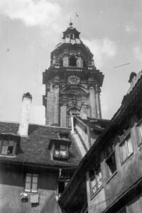 Diese historische Schwarz-Weiß Fotografie zeigt den Turm der Neubaukirche in Würzburg um das Jahr 1930. Der Turm steht im Zentrum des Bildes. Im Vordergrund sind die Fassaden von Wohnhäusern zu sehen.