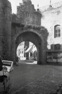 Dieses historische Schwarz-Weiß Foto zeigt das Nikolaustor in Würzburg um das Jahr 1930. Das alte Stadttor ist im Zentrum des Bildes zu sehen. An einem alten Gebäude rechts vom Tor hängen Werbeplakate. Im Torbogen sind die Häuser einer schmalen Gasse zu sehen. Am linken Bildrand ist jemand mit einem Kinderwagen unterwegs.