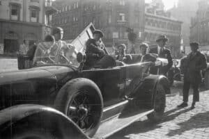 Dieses Foto zeigt ein altes Cabriolet aus den 1920er Jahren, das als Taxi an einem Platz in der alten Innenstadt von Würzburg steht. Im Taxi sitzen mehrere Männer, der Fahrer wartet neben dem Auto. Im Hintergrund sind alte Häuser und eine barocke Kirche zu sehen.